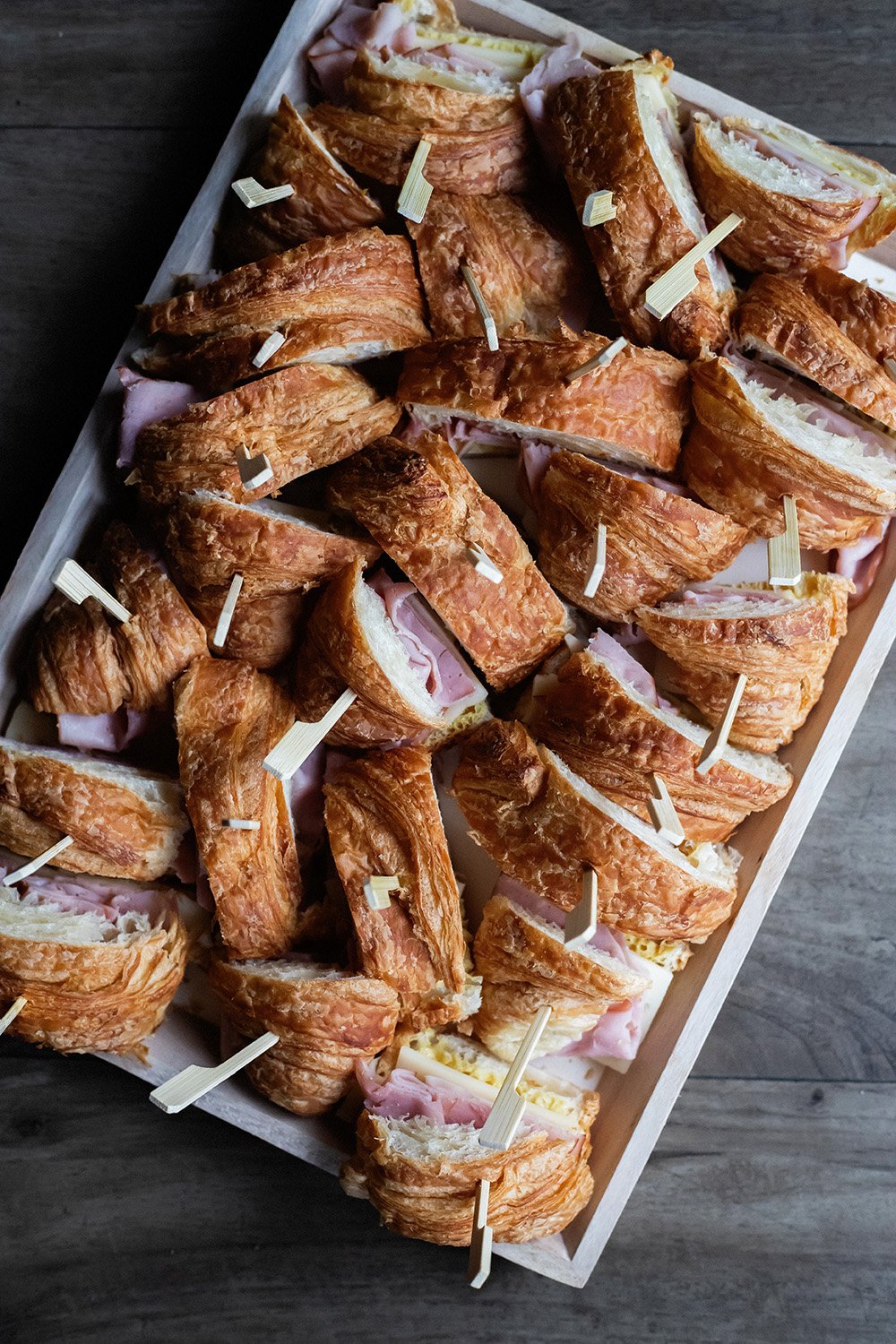 Assorted ham and cheese croissant sandwiches with skewers on a white tray.