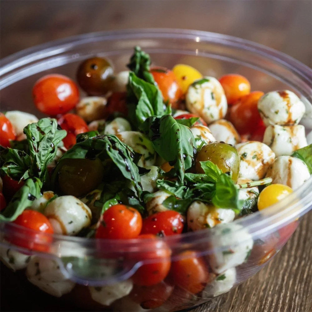 A plastic container filled with cherry tomatoes, fresh basil leaves, and mozzarella balls drizzled with balsamic glaze.