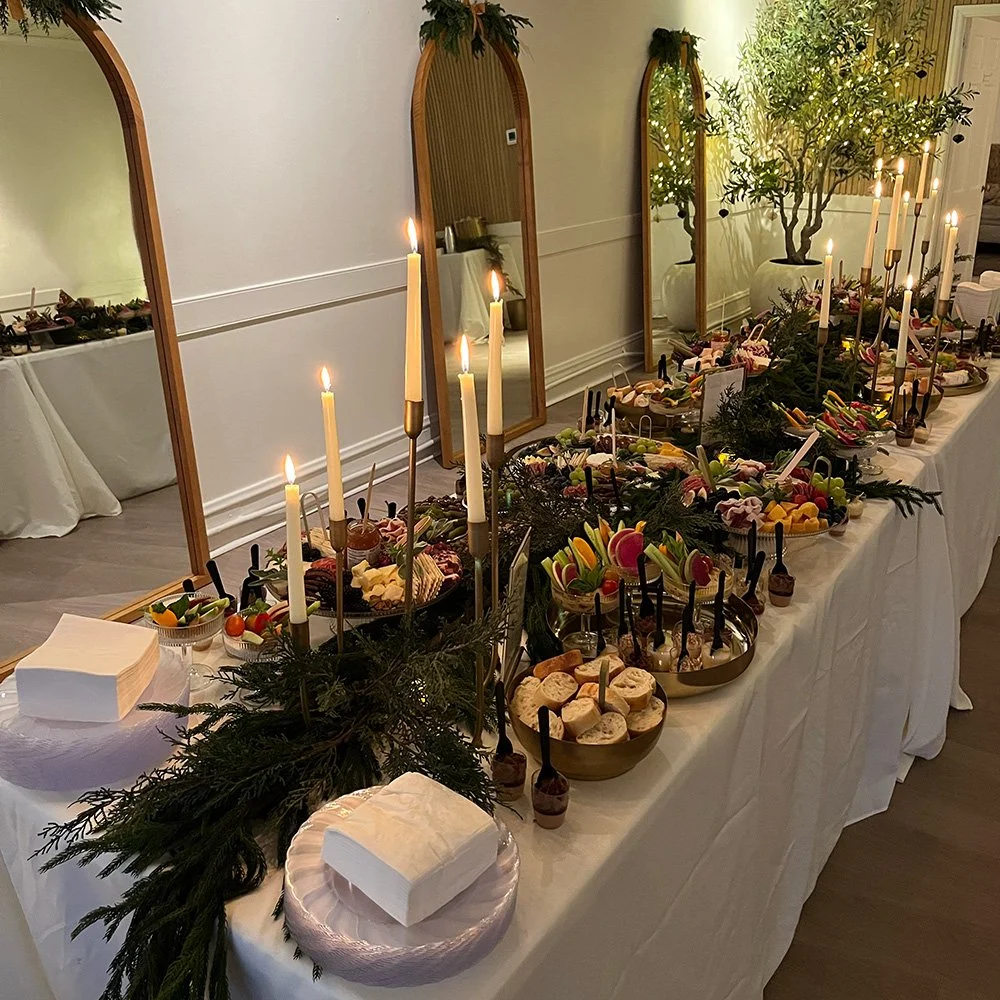 A long dinner table decorated for a celebration with a white tablecloth, candlesticks with lit candles, and plates of assorted candies, chocolates, and snacks. Large potted trees and tall standing mirrors are in the background.