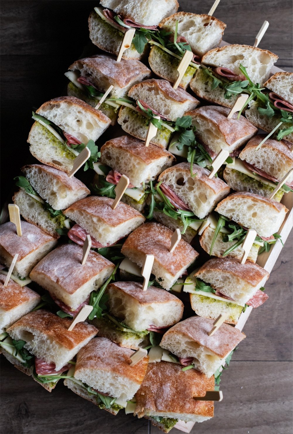 A tray of sliced sandwiches with crusty bread, ham, cheese, and arugula, held together with toothpick skewers.