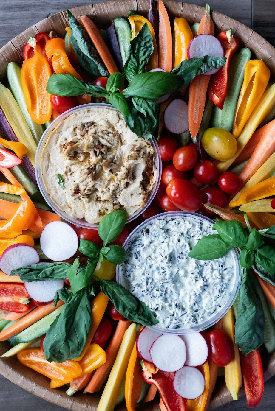 Colorful vegetable platter with cherry tomatoes, bell peppers, cucumbers, radishes, carrots, and leafy greens, accompanied by bowls of creamy dips topped with herbs.