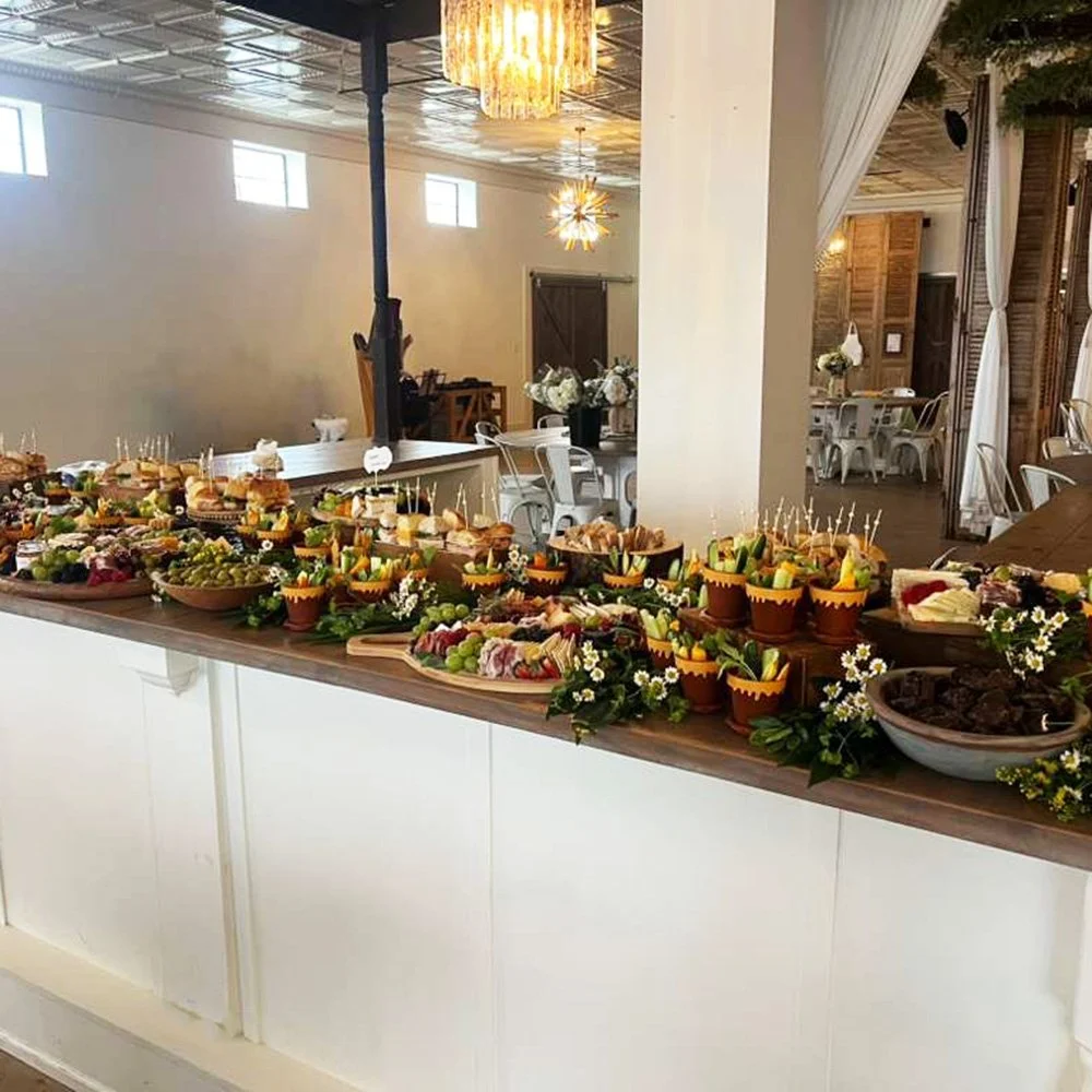 Decorative food display at a venue with assorted snacks, fruits, and small salads arranged on a wooden table, with white flowers and greenery for decoration.