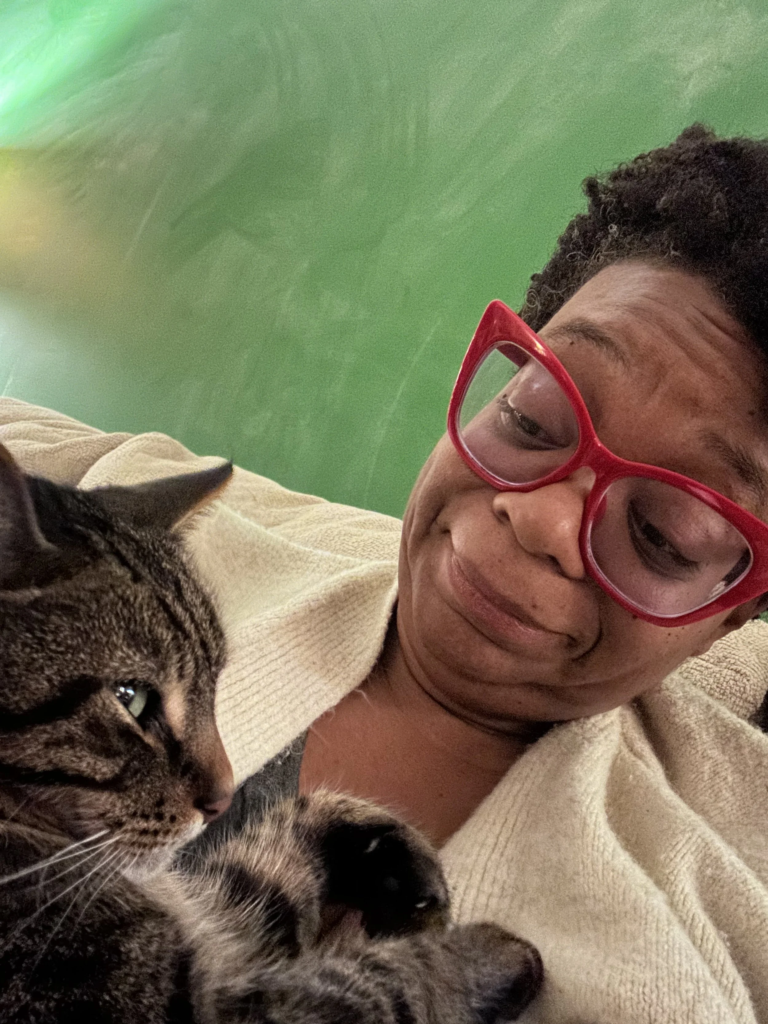 Chey Davis a Black woman with red catseye glasses and a white sweater smiling down at a tiger cat