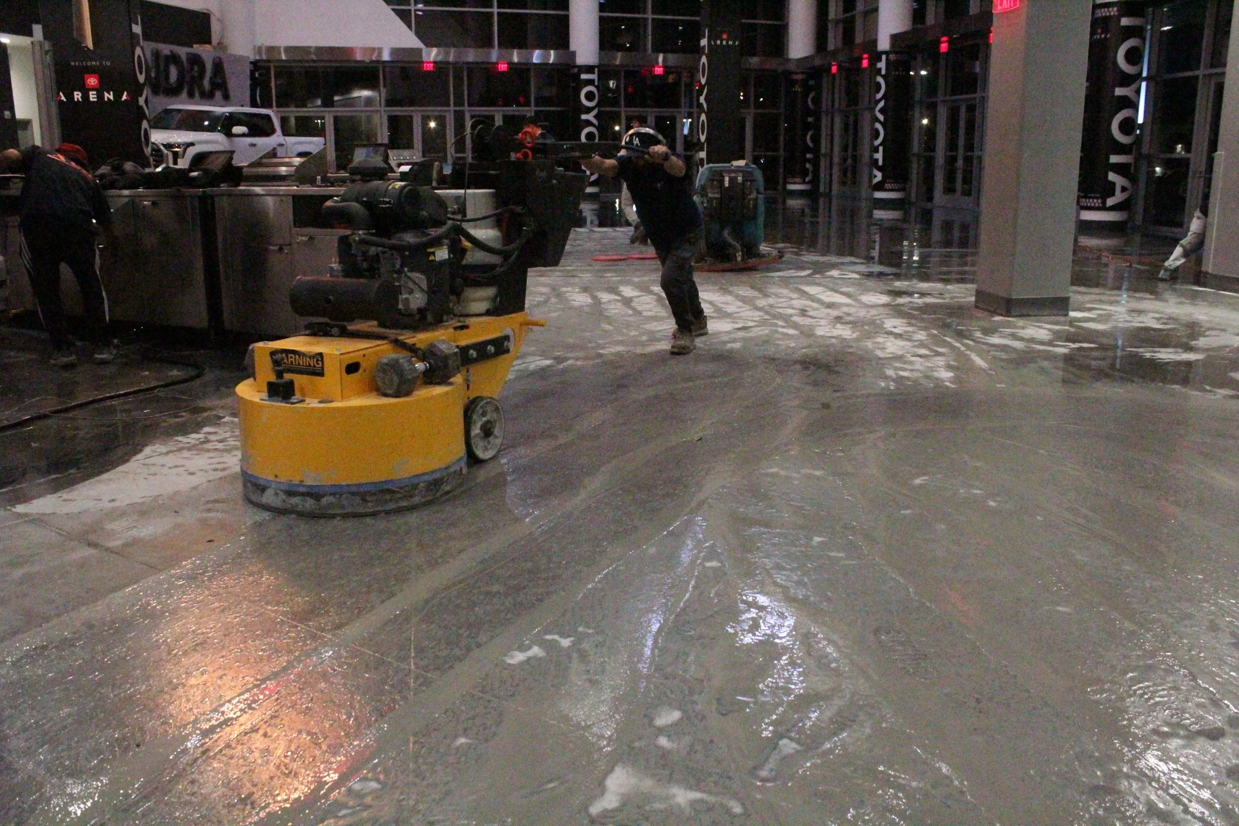 Worker buffing a large concrete floor with a large yellow machine. There is a solution on the ground that prevents dust and smooths the surface.