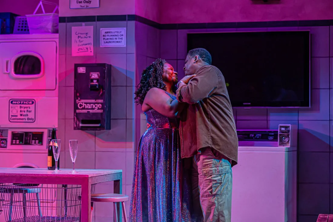A couple is dancing and holding each other closely in a brightly lit room with pink and purple lights, surrounded by laundry machines and a television.