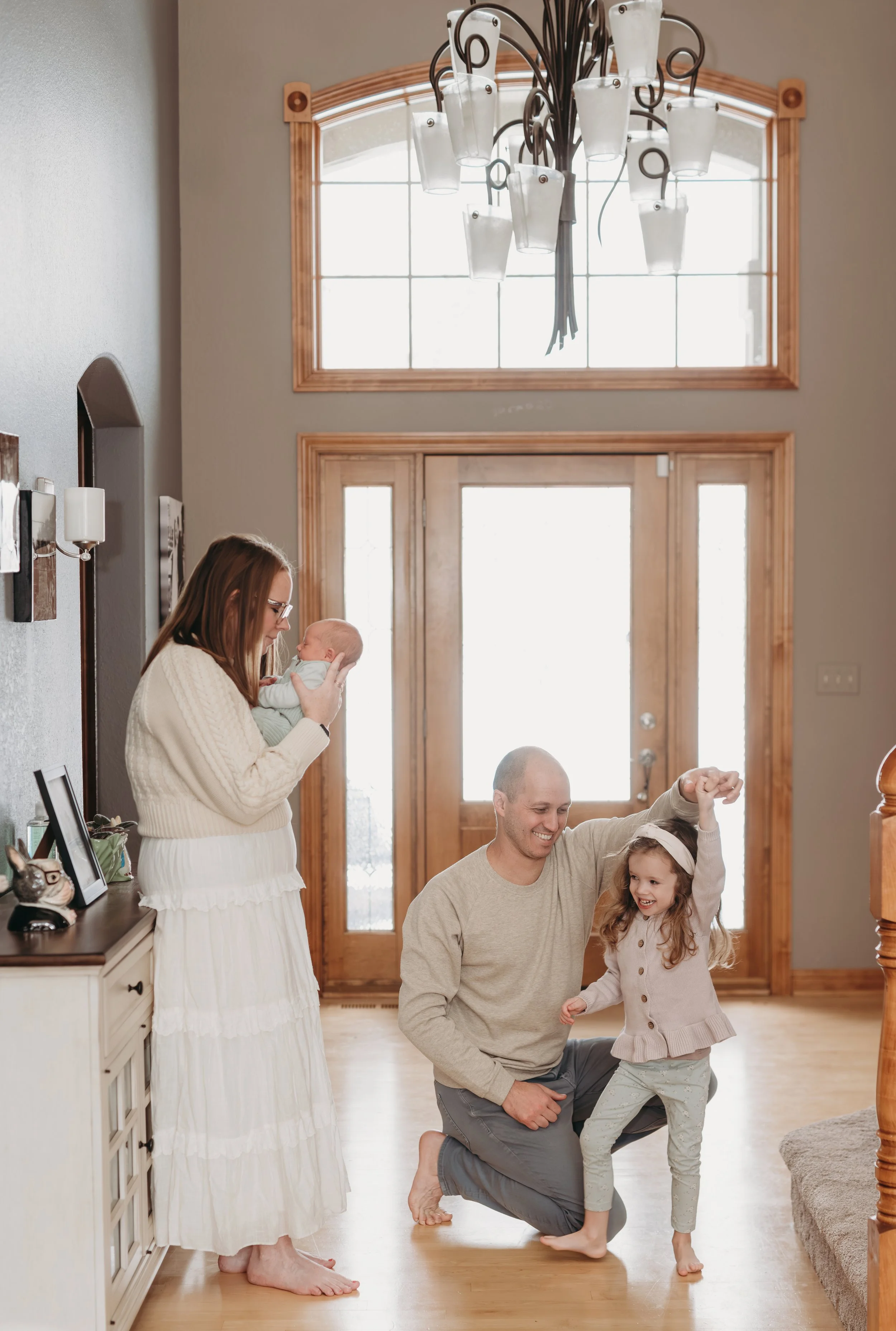 A family in a warmly lit foyer, with a woman holding a baby, a man kneeling and playing with a young girl, near a wooden door with glass panels and a large window above.