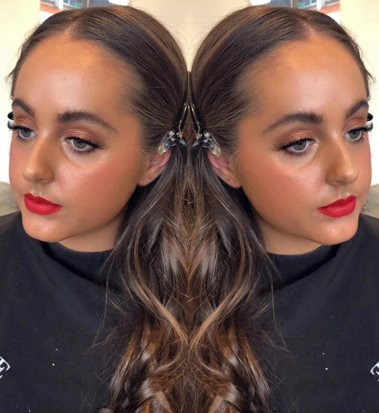 A woman with long wavy brown hair, wearing red lipstick and a black top, against a neutral background.