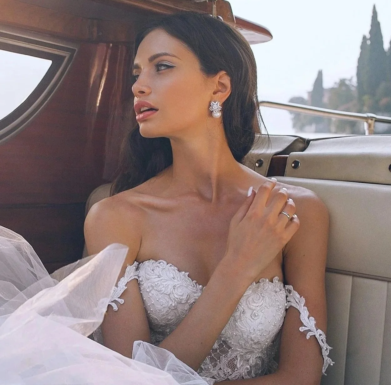 A woman in a wedding dress with lace details and pearl earrings sitting inside a vintage car with wooden interior, overlooking a scenic background with trees.