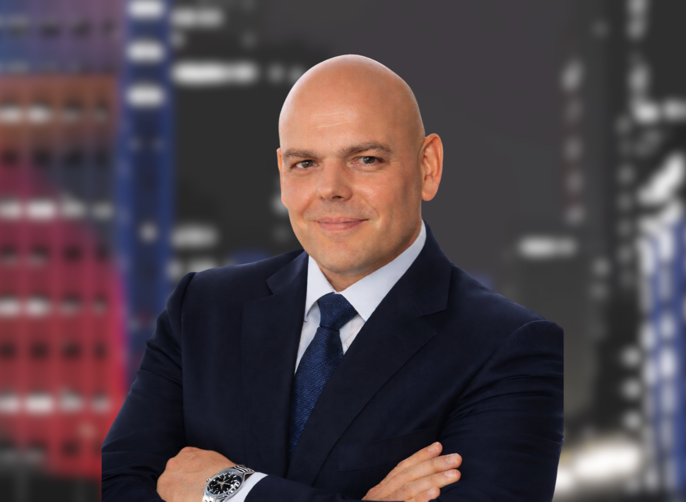 Headshot of a bald man wearing a suit and a red and white striped shirt and tie, with a cityscape background.
