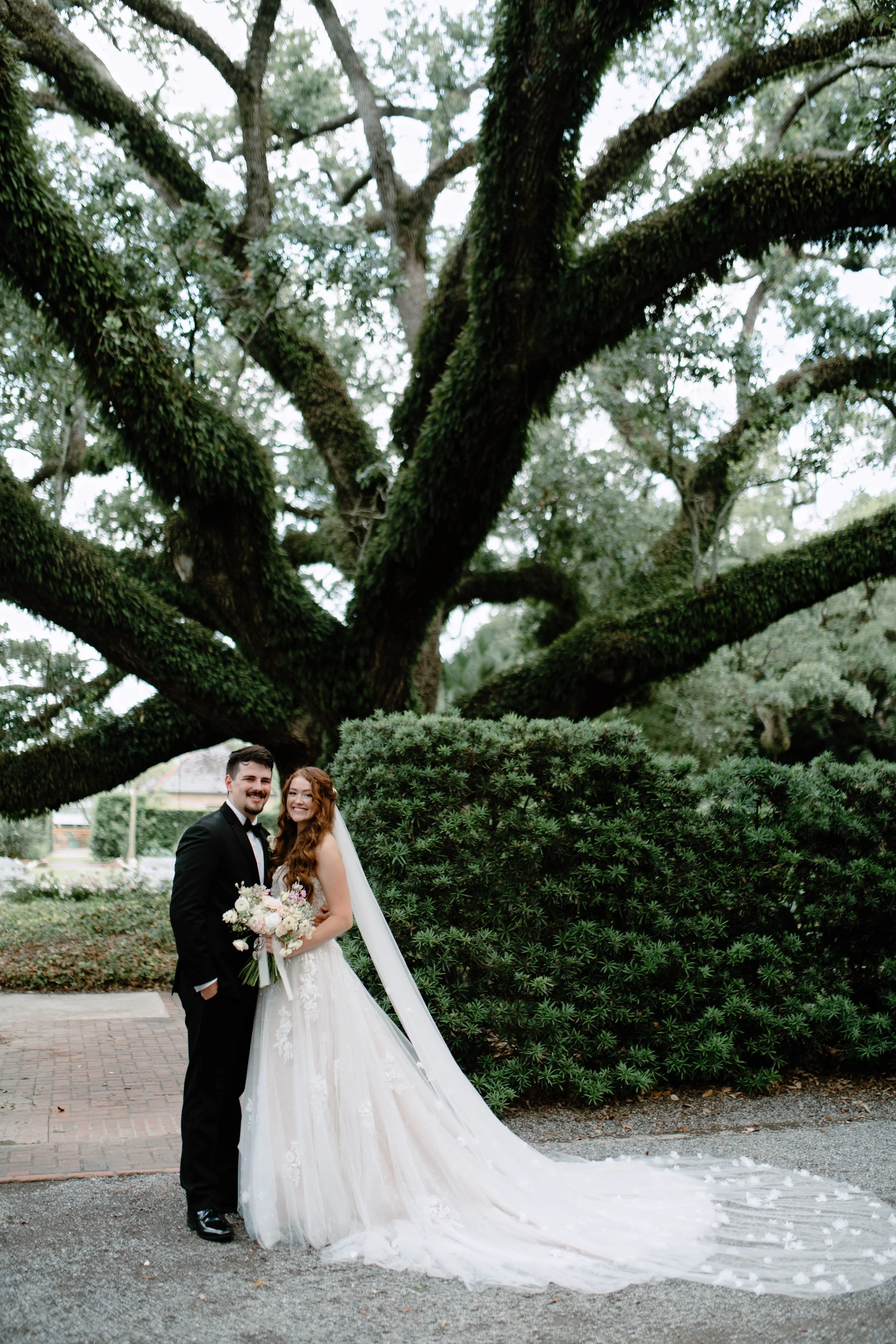 A bride and groom smiling outdoors under a large, moss-covered tree on a cloudy day. The bride wears a white wedding gown with a long train, and the groom wears a black tuxedo. The bride holds a bouquet of white and pink flowers. There are bushes and a brick pathway in the background.