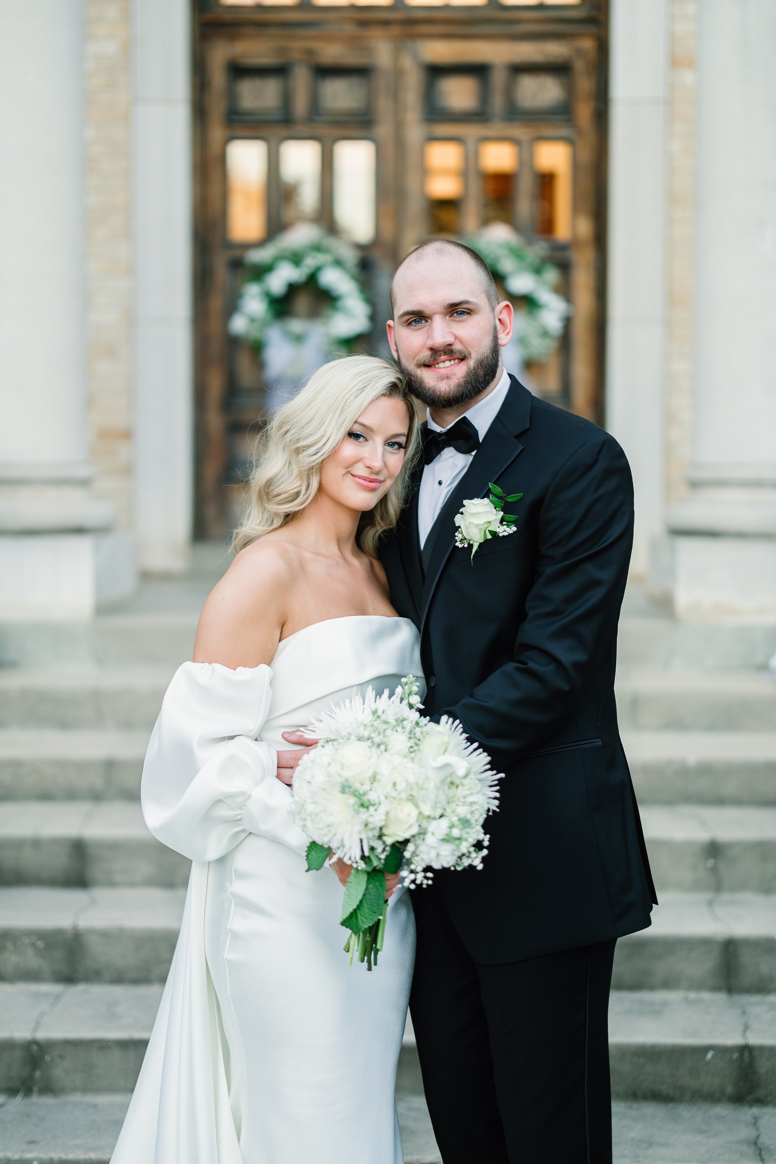 A bride and groom standing side by side on the steps of a church, smiling at the camera. The bride is holding a bouquet of white flowers and wearing an off-the-shoulder white wedding gown. The groom, dressed in a black tuxedo with a white shirt and black bow tie, has a white flower boutonniere on his lapel. The church entrance behind them is decorated with floral arrangements.