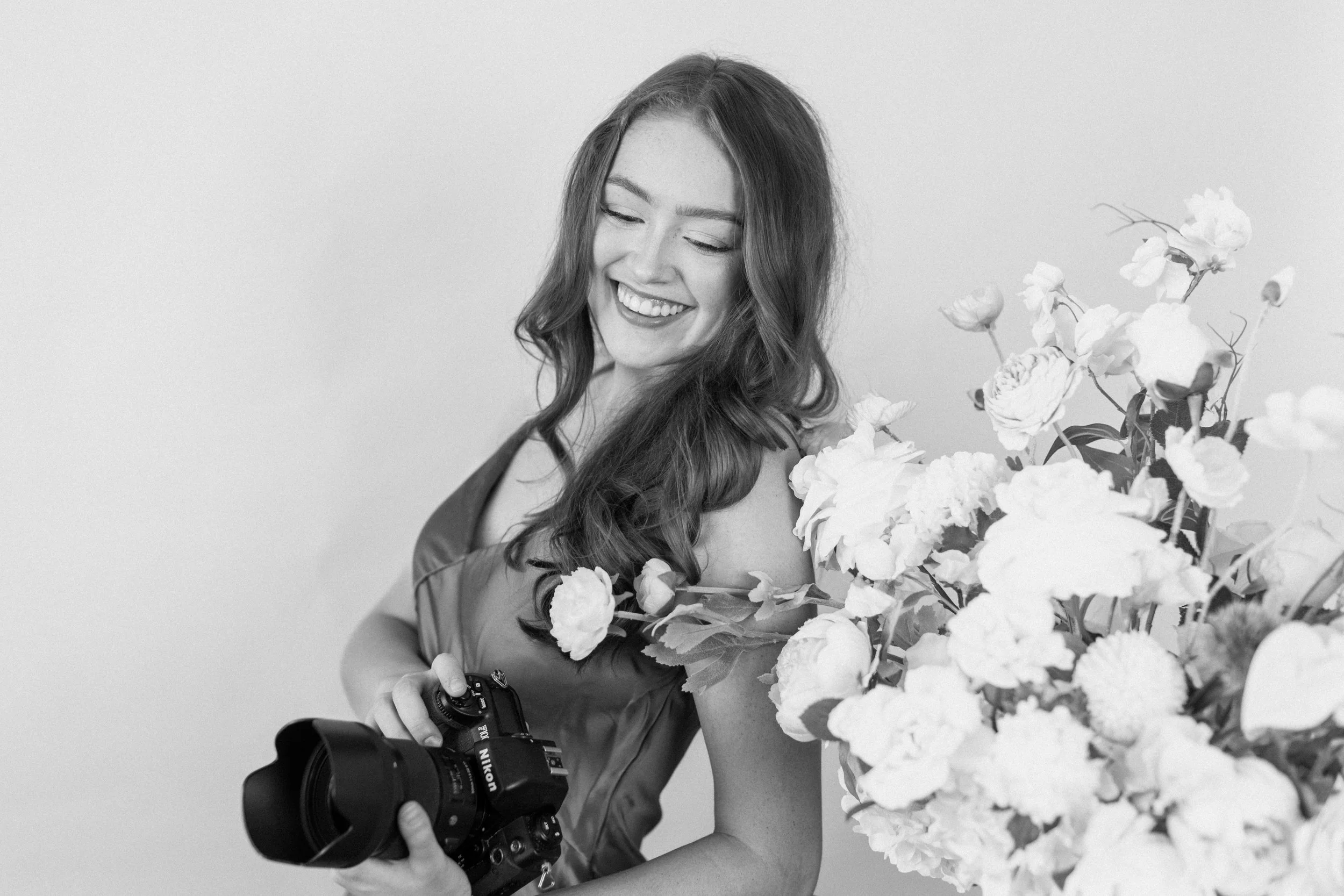 A smiling woman with long, wavy hair holds a professional camera and stands beside a large bouquet of flowers, against a plain light background.