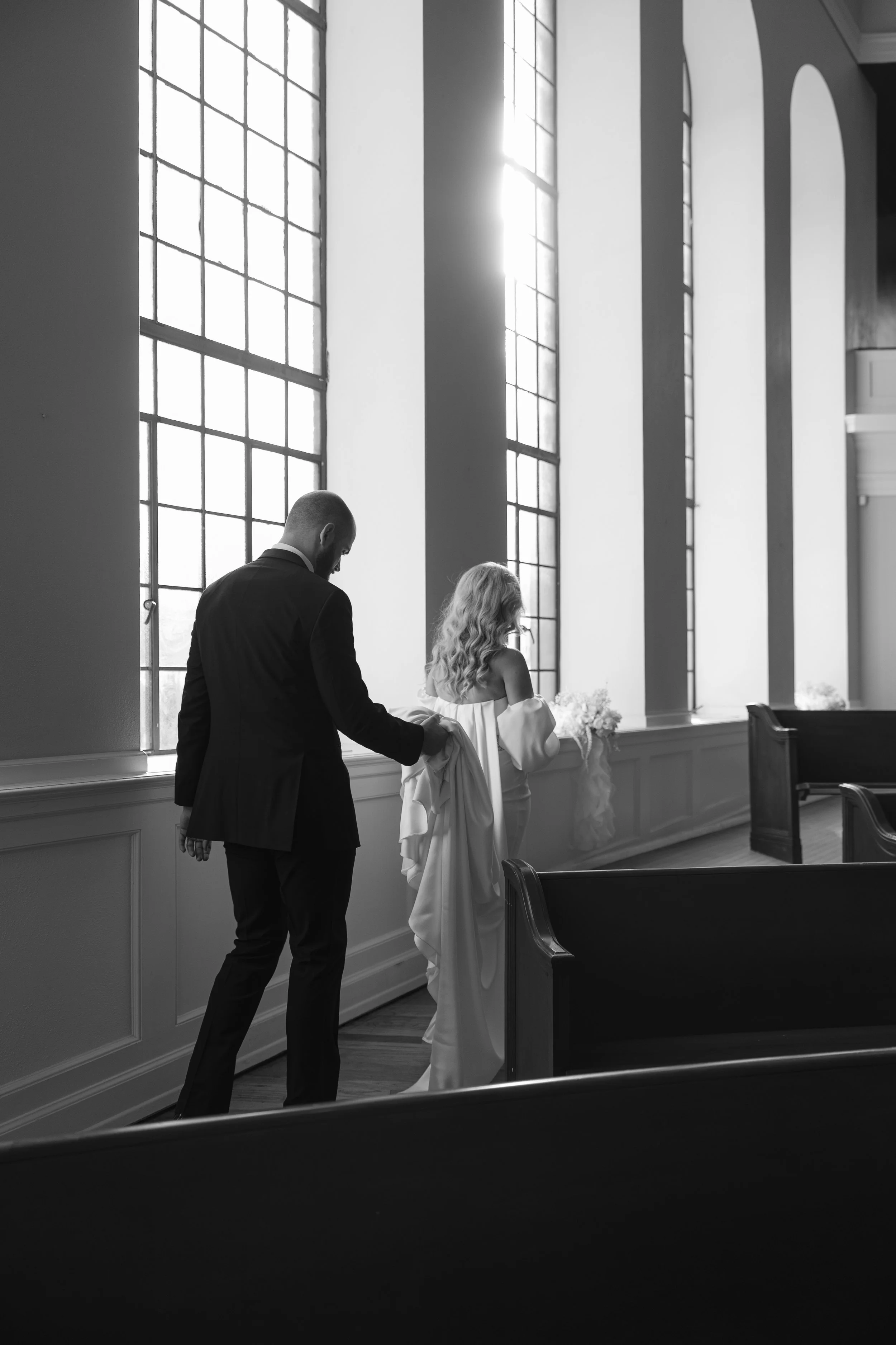 A black-and-white photo of a bride and groom standing inside a church with large stained glass windows, the bride holding a bouquet and the groom assisting her with her wedding dress.