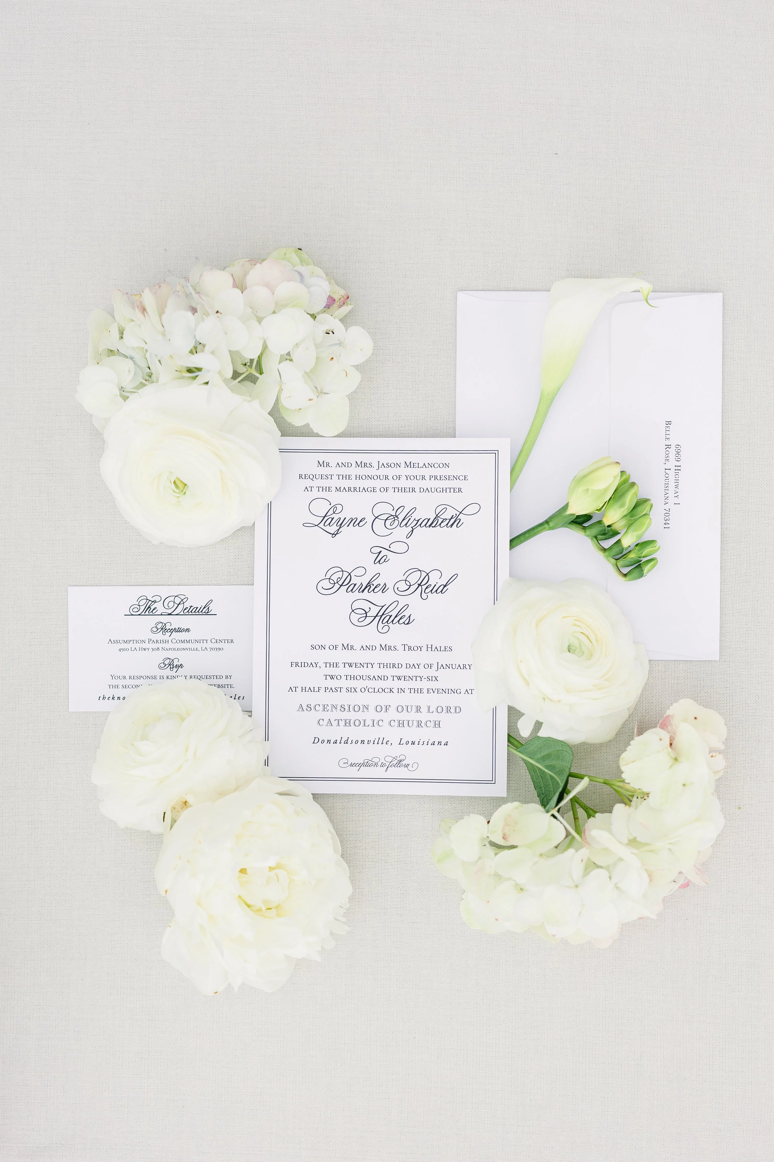 Wedding invitation surrounded by white flowers and an envelope on a light fabric surface.