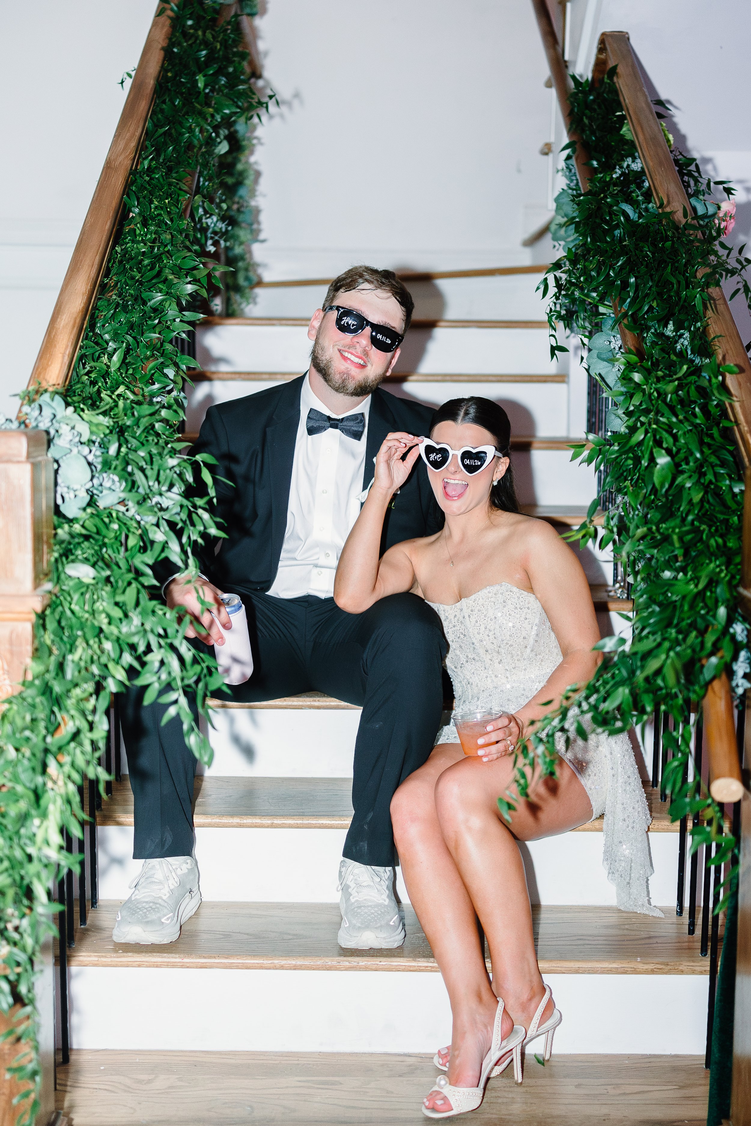 A group of three people, two men and one woman, sitting on a staircase decorated with green foliage. They are dressed in formal attire, with the man in a tuxedo and the woman in a white dress. All are wearing sunglasses with white frames and black writing, and they are smiling and making playful poses. The woman is holding a drink and is wearing high-heeled shoes.