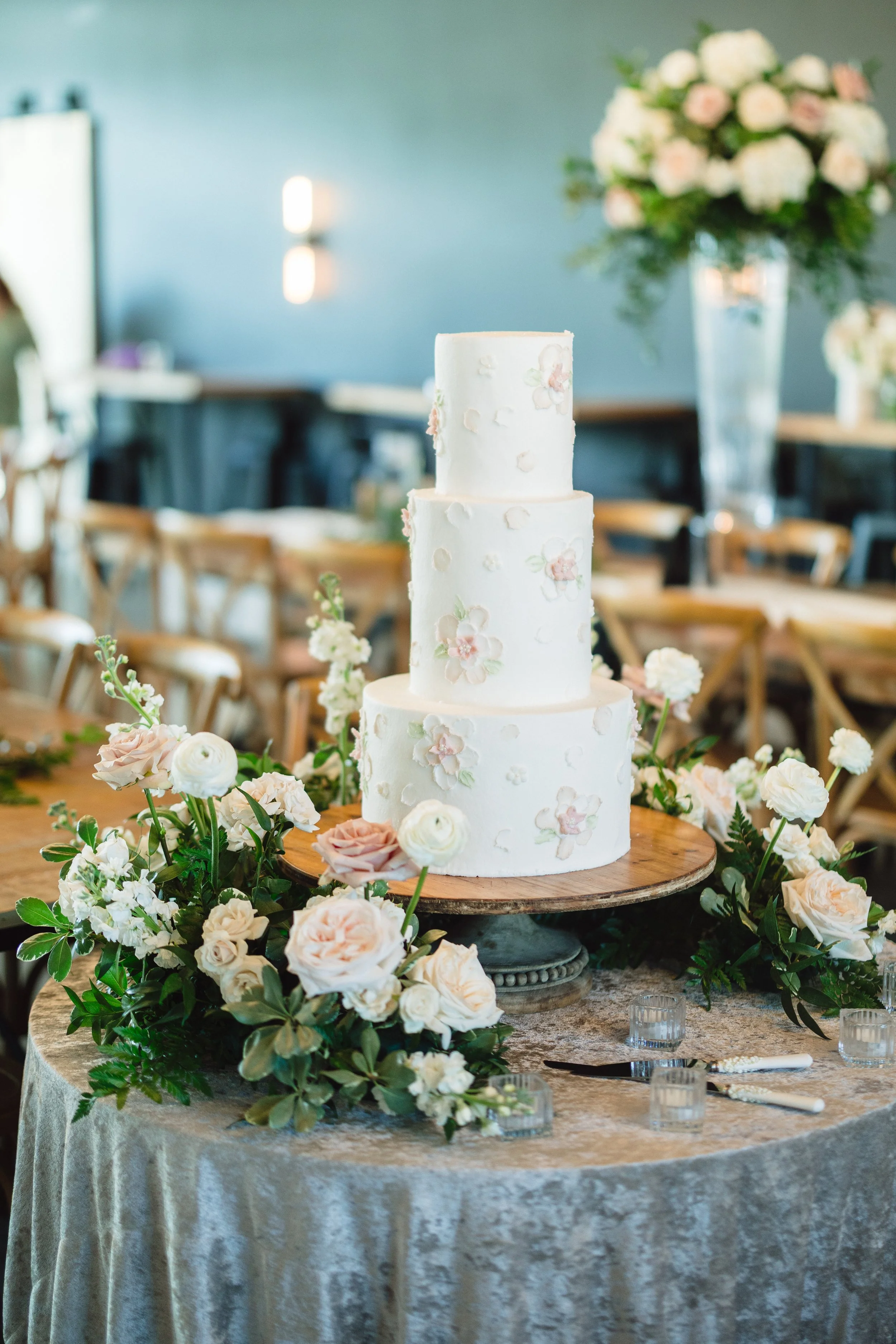 A three-tiered wedding cake decorated with pastel pink and white flowers, surrounded by a lush arrangement of white and blush-colored roses and greenery on a round table with a lace tablecloth. In the background, there are chairs and a large floral arrangement in a tall vase.