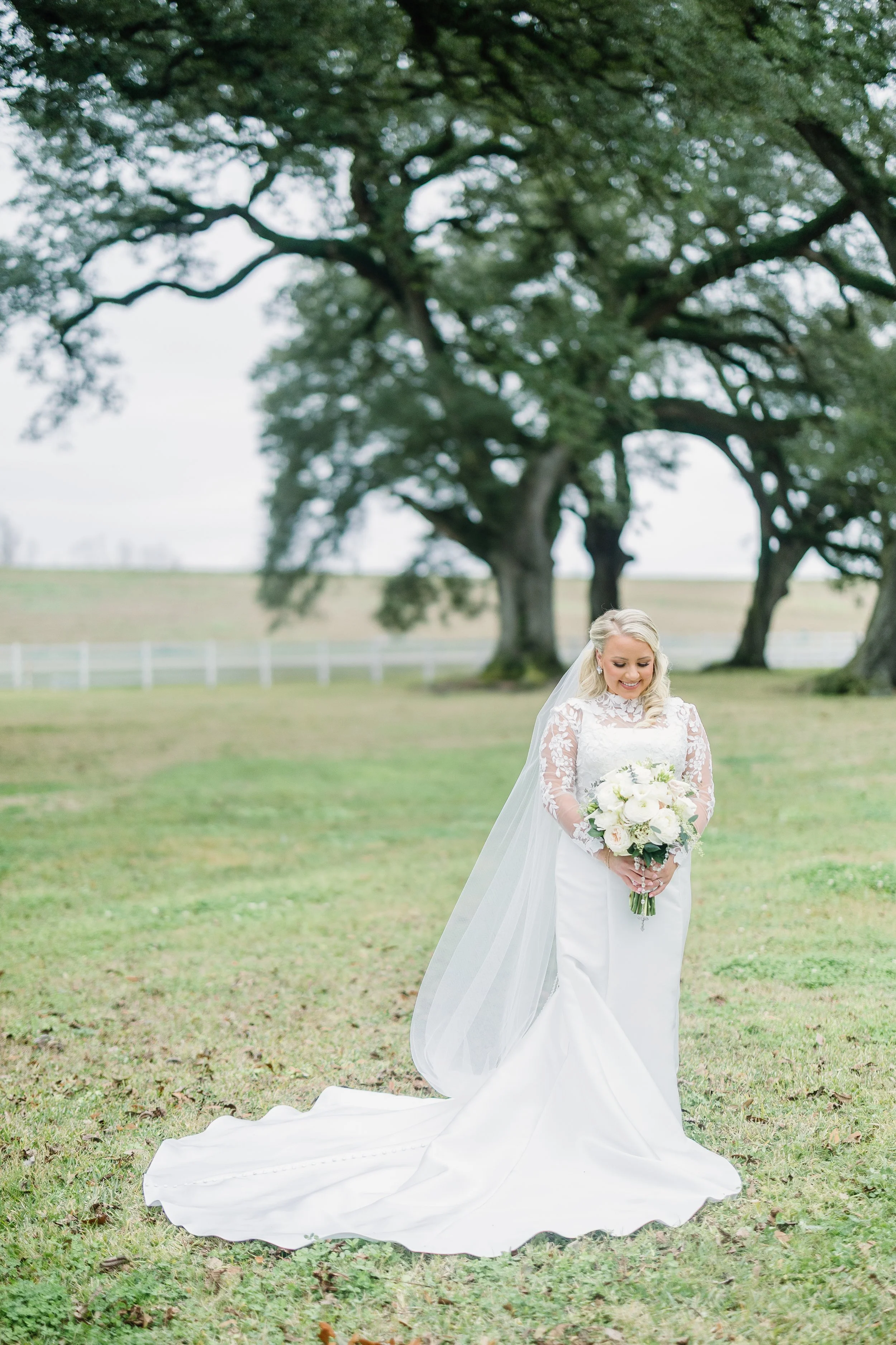 Bride in a white wedding dress holding a bouquet of white flowers, standing outdoors on grass with large trees in the background.