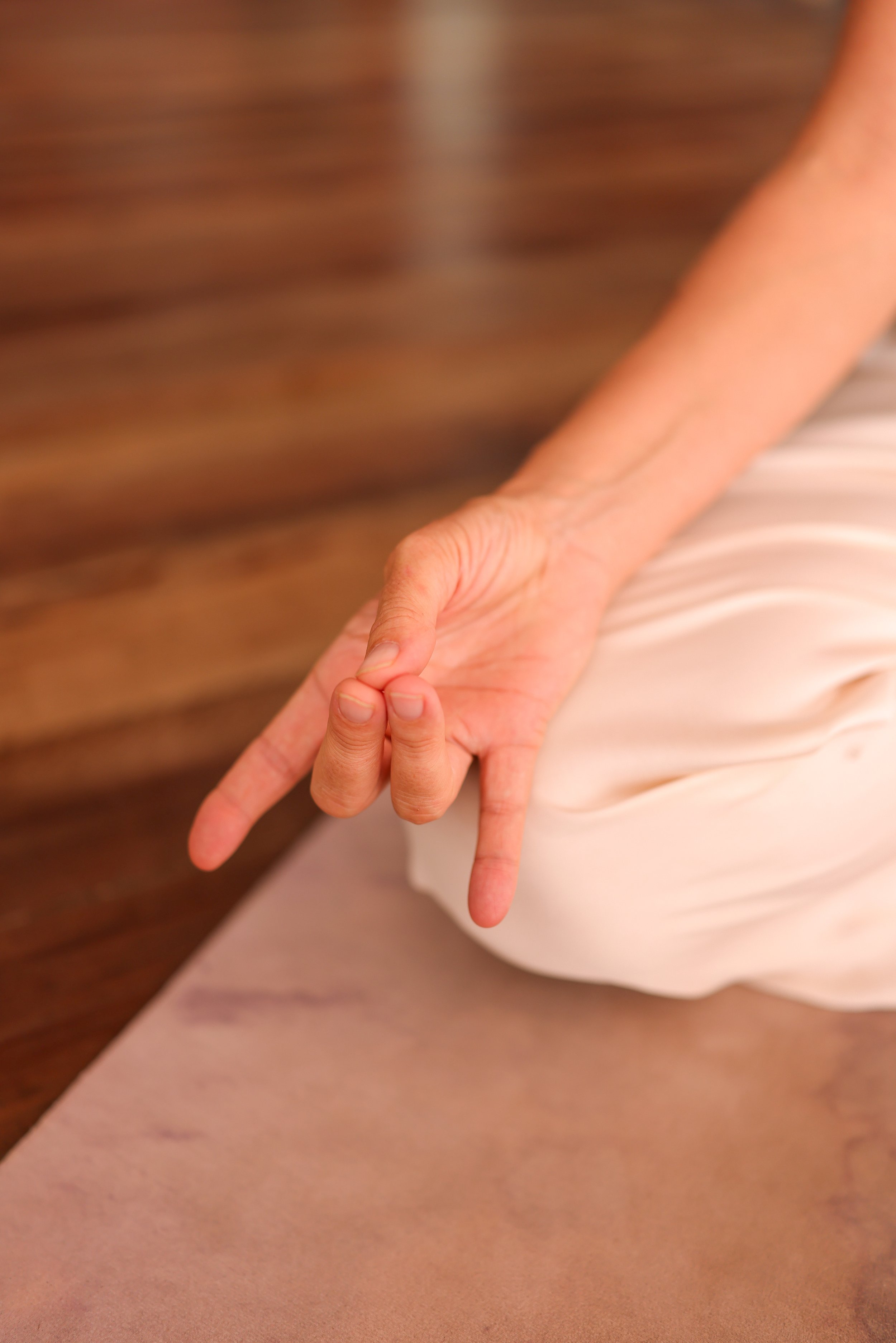 Close-up of a person making a peace sign with their fingers, wearing a cream-colored dress, with a blurred wooden background.