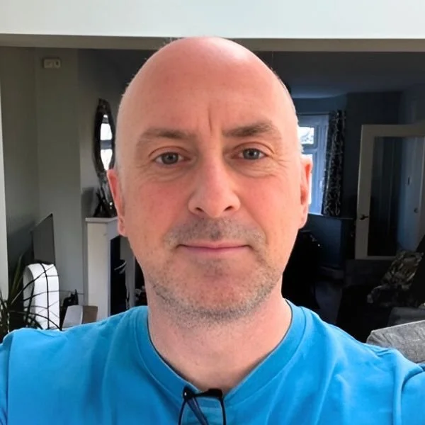 A middle-aged bald man with blue eyes and light facial hair, wearing a bright blue shirt, standing indoors in a home setting with a living room background.