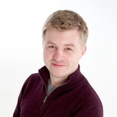 A young man with short light brown hair wearing a dark maroon zip-up sweater, smiling slightly, against a plain white background.