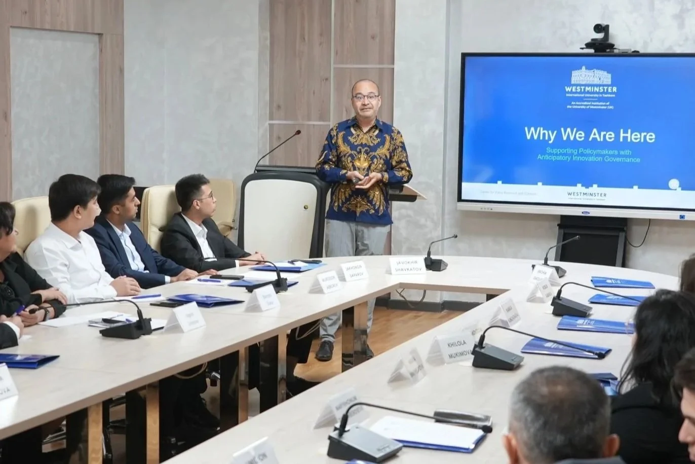 A man giving a presentation at a conference room with seated attendees and a large screen displaying the title 'Why We Are Here' at Westminster, supporting policymakers with anticipatory innovation governance.