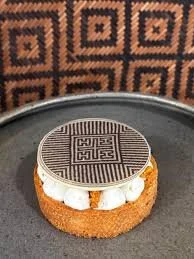 Dessert with a decorative circular chocolate piece on top, layered with cream, and a biscuit or cake base, served on a round black plate.