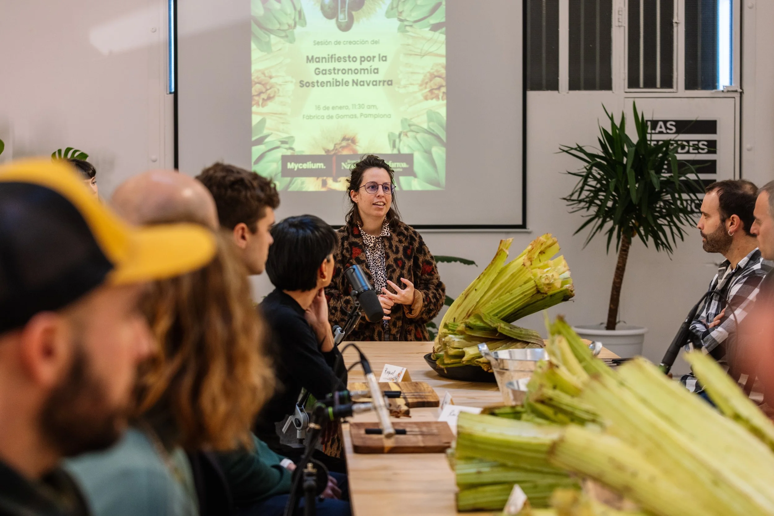 A woman speaking at a conference or meeting, standing in front of a projected presentation slide titled 'Manifesto for Sustainable Gastronomy Navarre', with a conference table of people and bananas on it.
