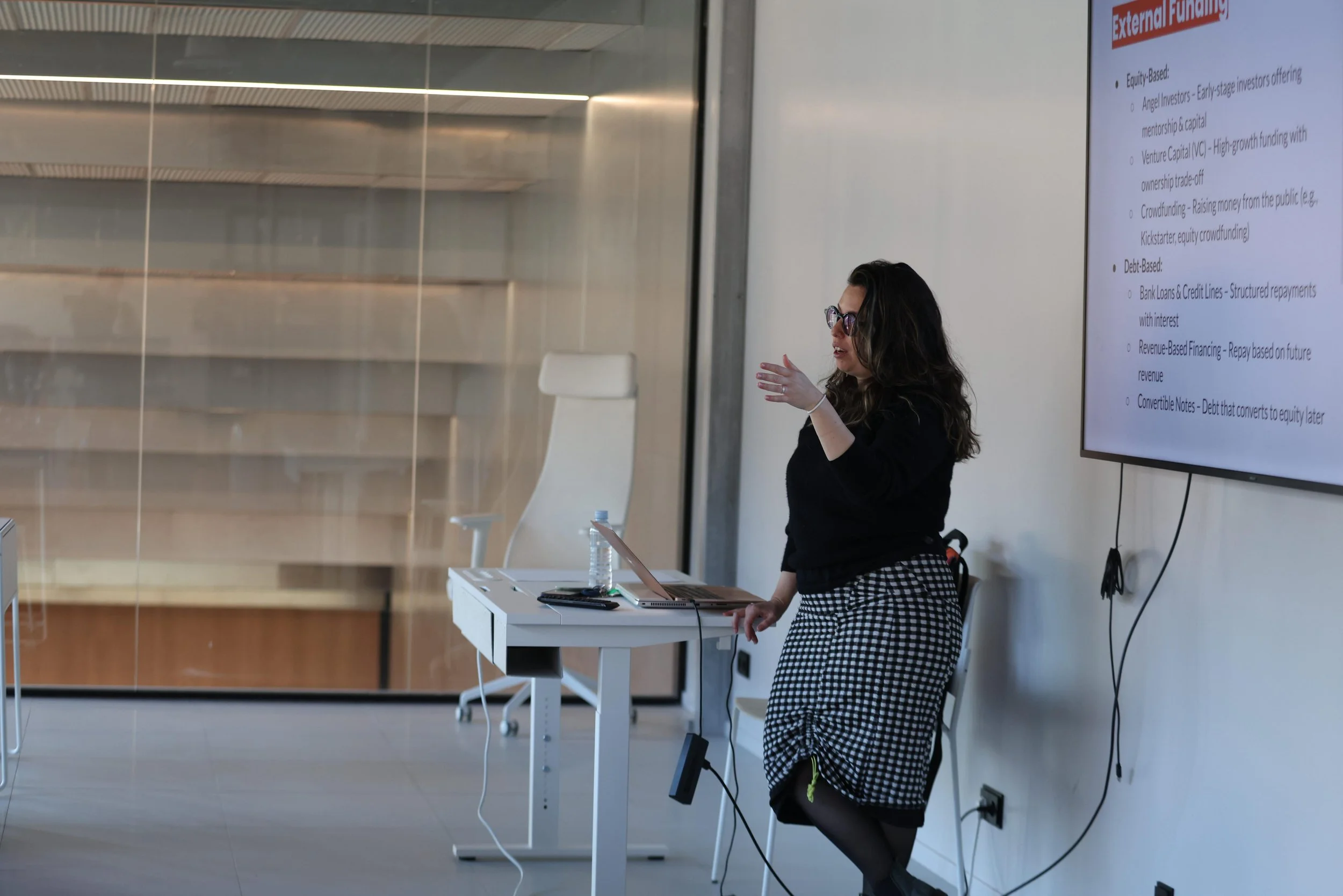A woman giving a presentation in an office or conference room. She is standing next to a large screen displaying a slide with bulleted text. She wears glasses, a black top, and checkered pants, and has long dark hair. There is a laptop, a water bottle, and some papers on a white table in front of her.