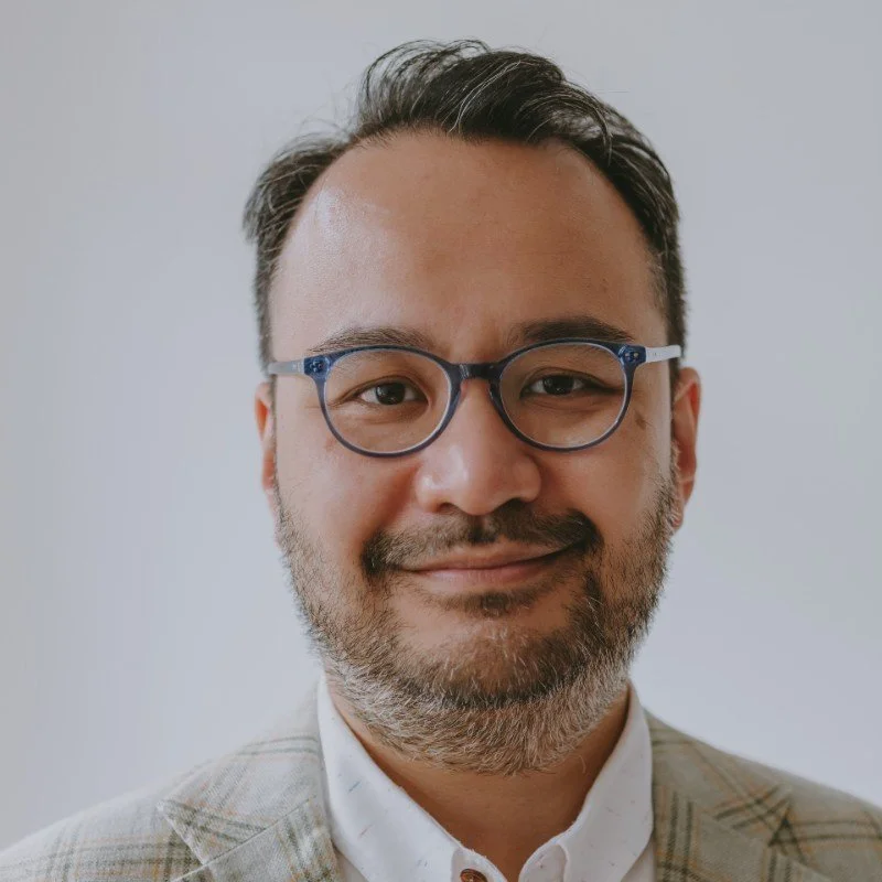 A man with glasses and a beard smiling, wearing a light-colored blazer over a white shirt.