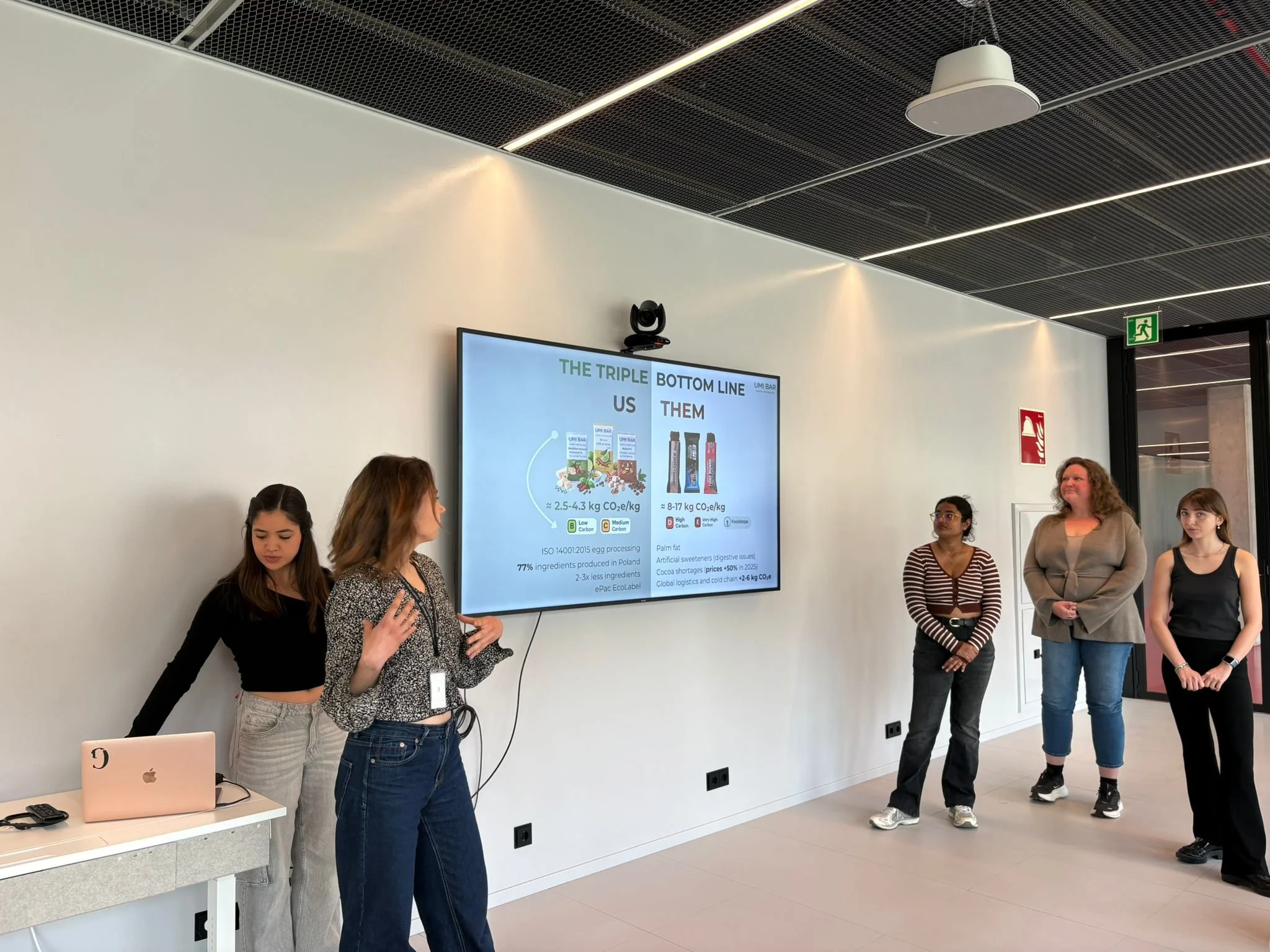 A group of people standing in front of a large screen displaying a presentation in a modern conference room with white walls and black ceiling panels.