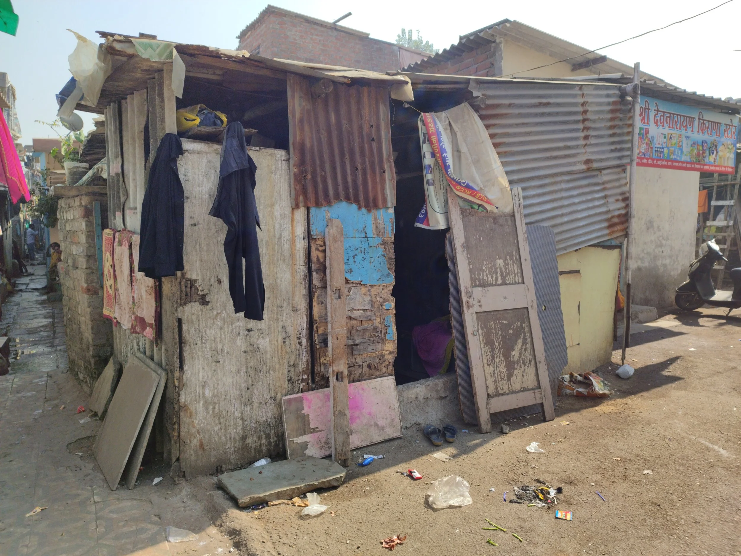An informal shelter made of bits of wood, sheets of rusty corrugated iron and other materials. There is not a proper front door and there are large gaps in the wall. It is very dark inside. The occupier has left their shoes outside the entrance.