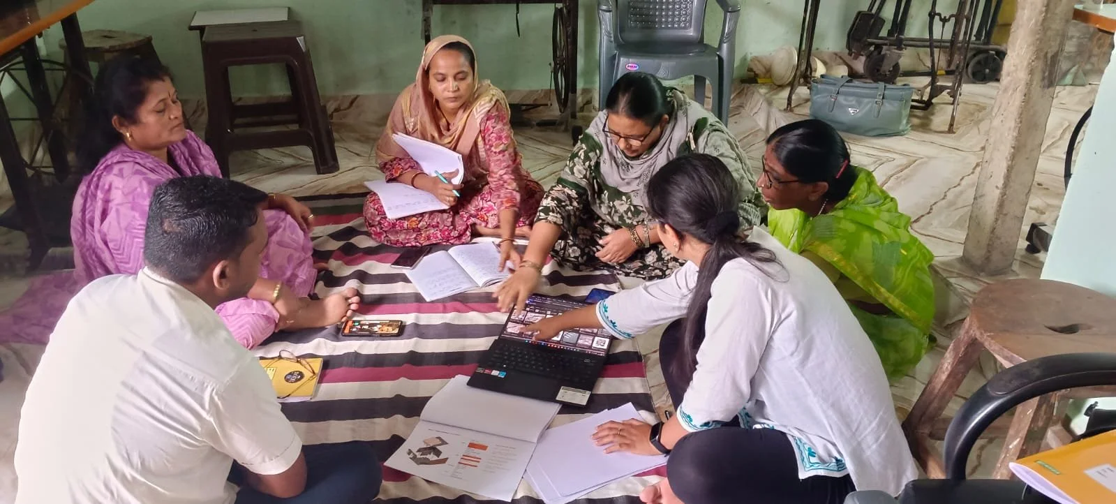 five women and one man sit in a circle on the floor, in discussion around a computer and papers