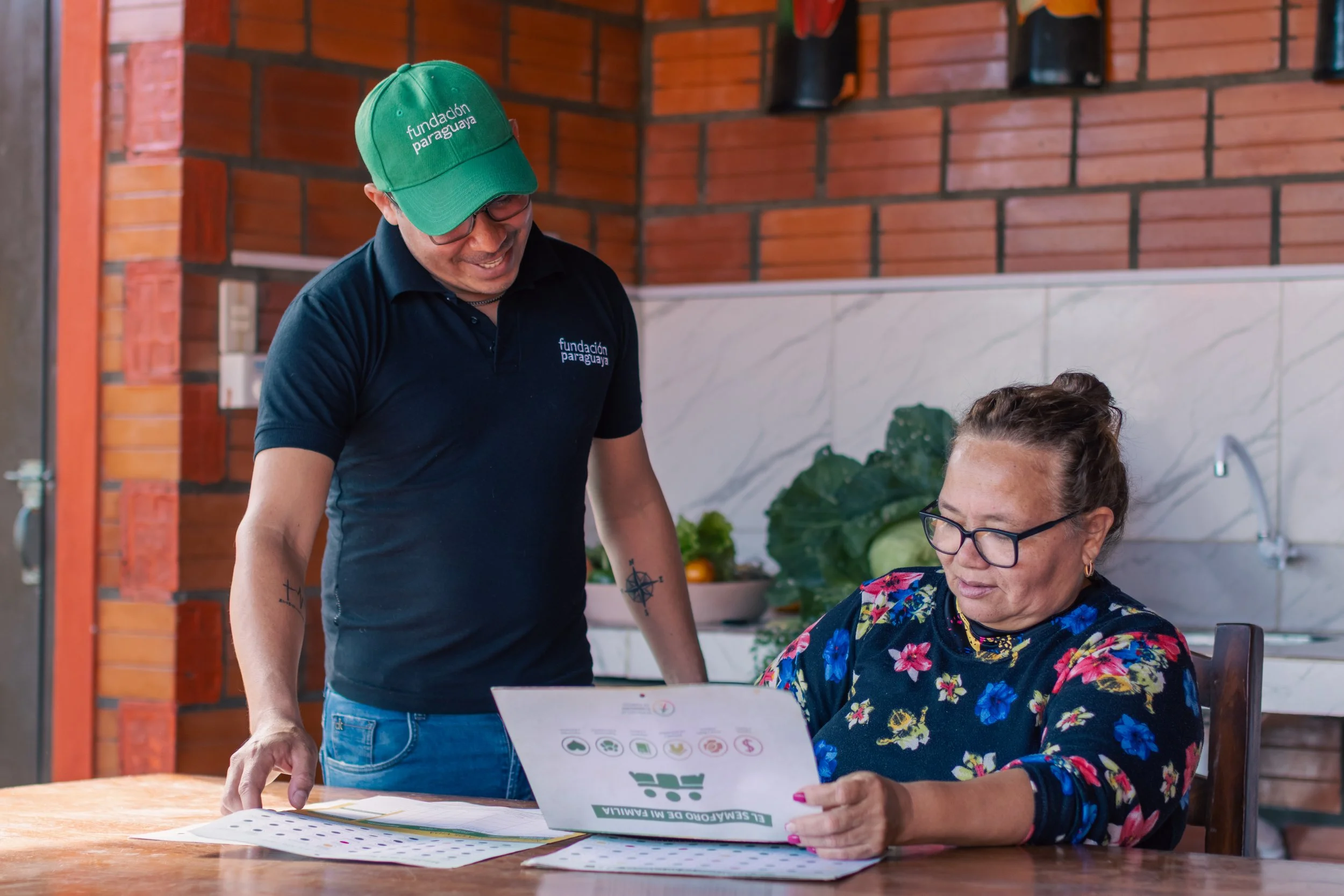 Middle-aged woman sitting at kitchen table looking at Poverty Spotlight documents. A man wearing a green Fundcion Paraguaya baseball cap stands next to her smiling while looking at what she is pointing at on the documents.