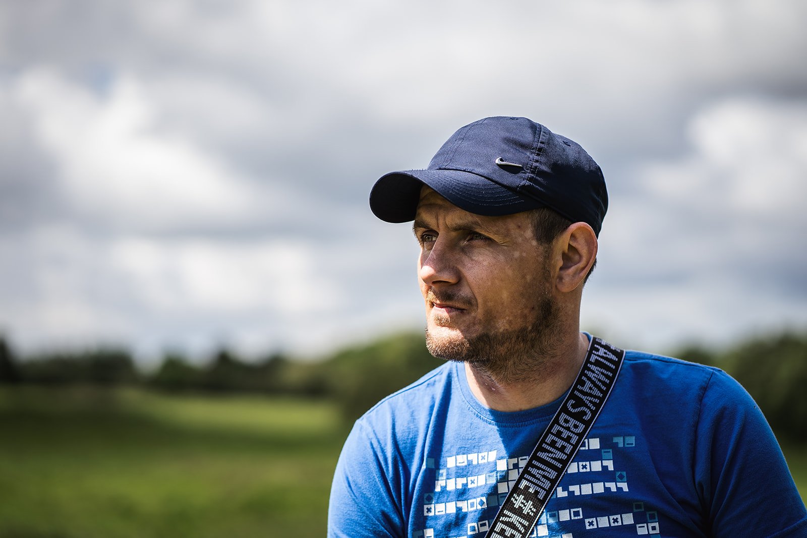 Man sitting in the countryside, wearing a blue t-shirt and dark blue cap.