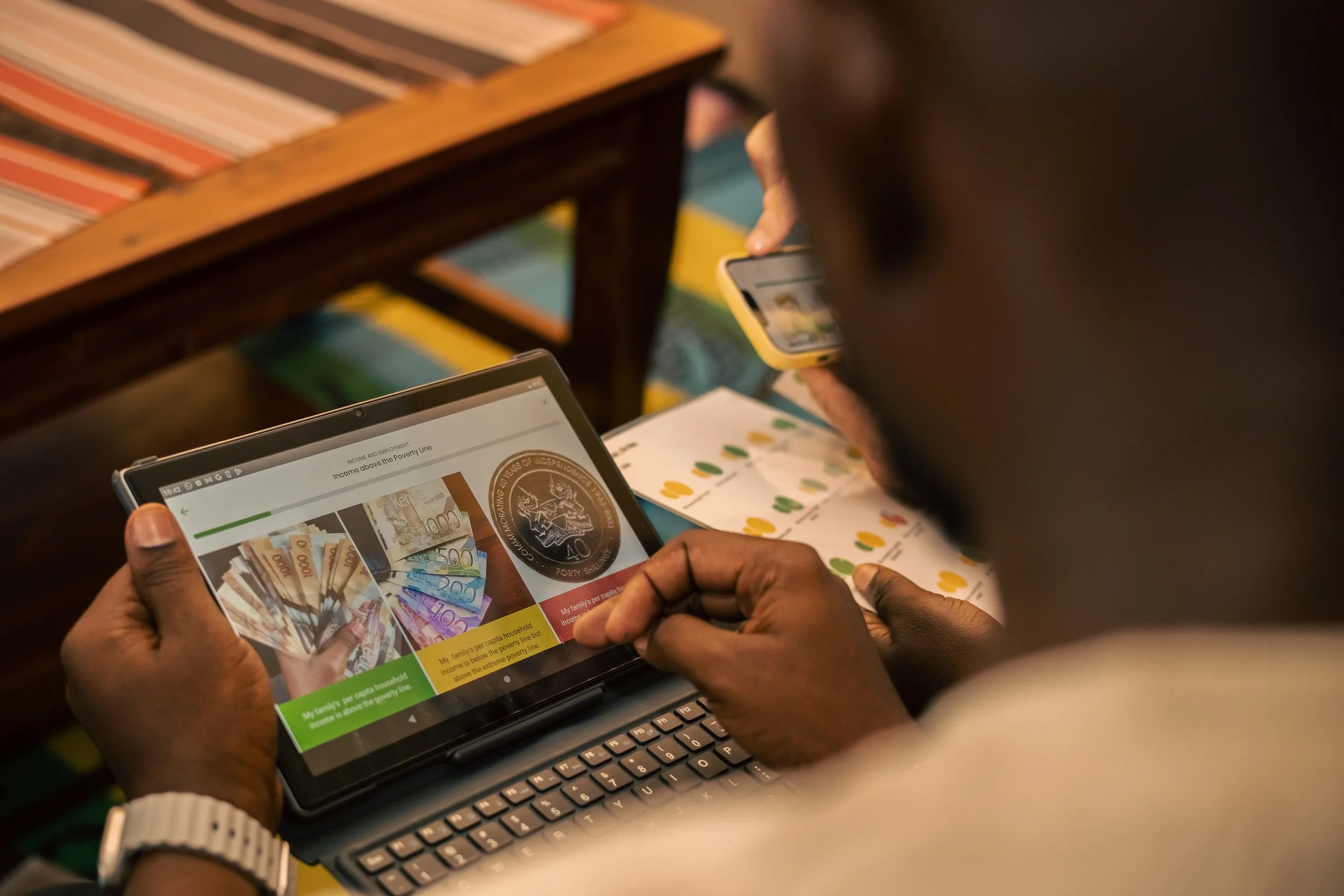 Image showing three people’s hand’s - one is holding a small computer with the Poverty Spotlight programme on screen, one is holding a smartphone with a different programme image and one is holding a printed document.