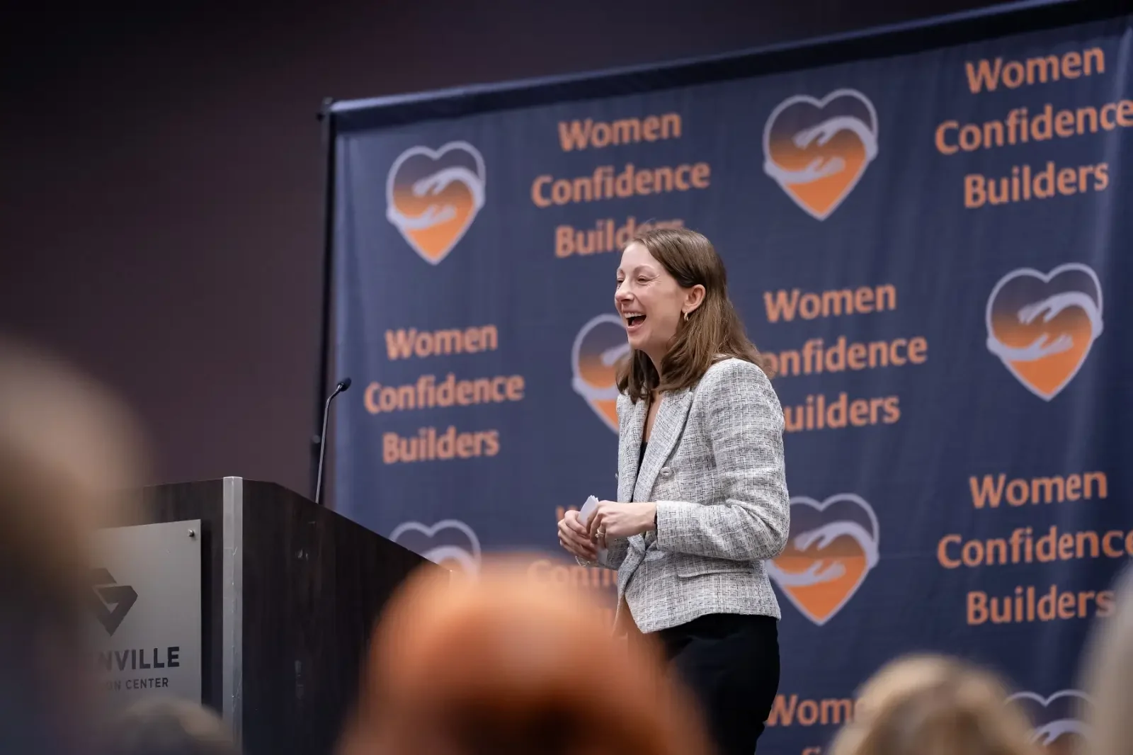 Speaker presents on stage in front of a Women Confidence Builders backdrop during a professional event.