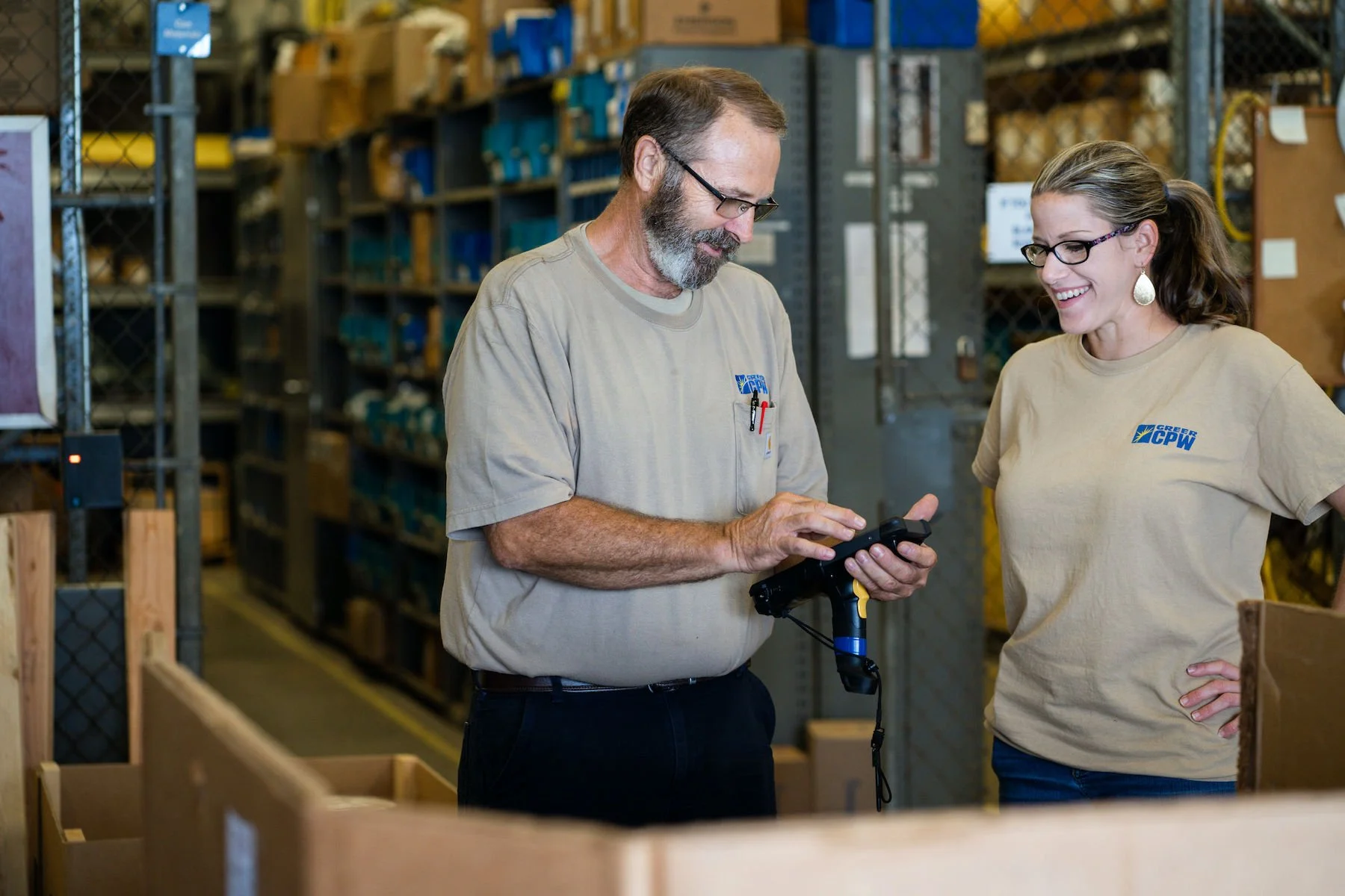 Two skilled trades team members smile while reviewing a mobile device in a warehouse work area.