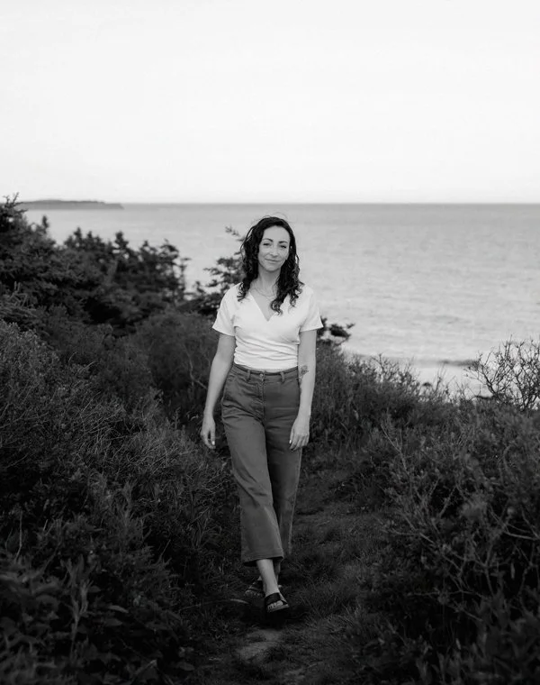 Black & white photo of a woman walking a path between some shrubbery. She has curly hair and a white shirt.