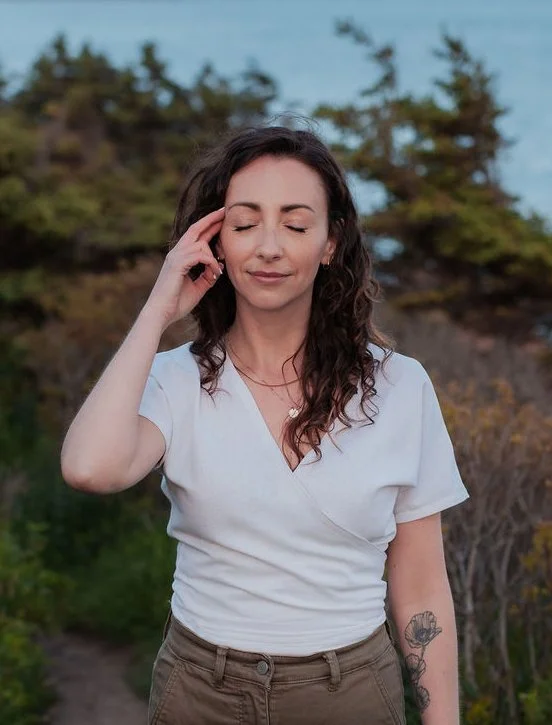 Image of woman with curly brown hair tapping beside her eye with two fingers. She wears a white short sleeved shirt and olive green pants.