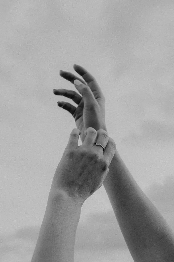 Black and white image of hands reaching into the sky. There is a dainty ring on the middle finger of the left hand.