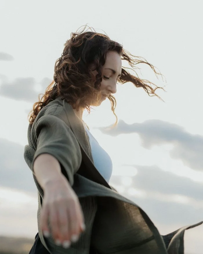 Woman with curly hair is backlit by the sun. She moves her arms and her sweater is billowing in the wind.