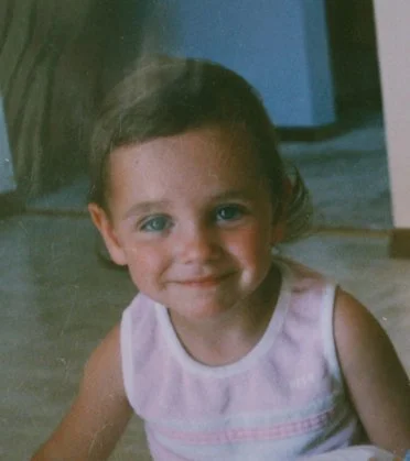Image of a young girl with a sweet smile on her face; she wears a pale pink tank top with white trim