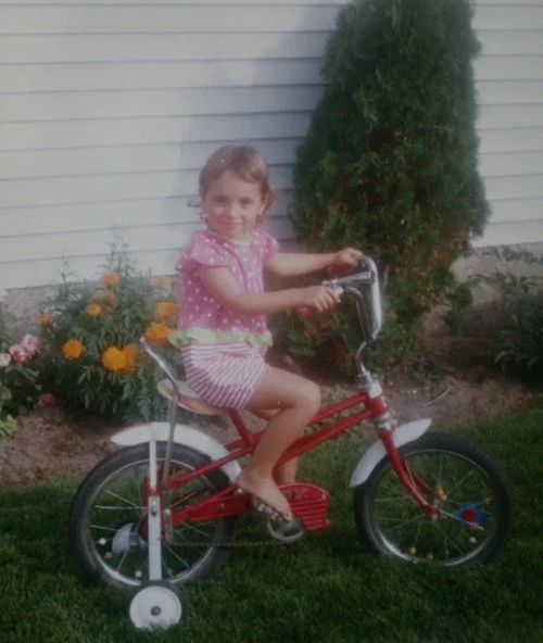 Photograph of a young girl in a pink polka dot shirt and striped shorts on a red bicycle with training wheels; there is green grass, flowers, and white house siding in the background
