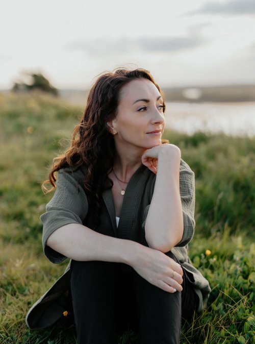 Woman with curly brown hair is looking pensively off-camera. She wears a green sweater and black pants.