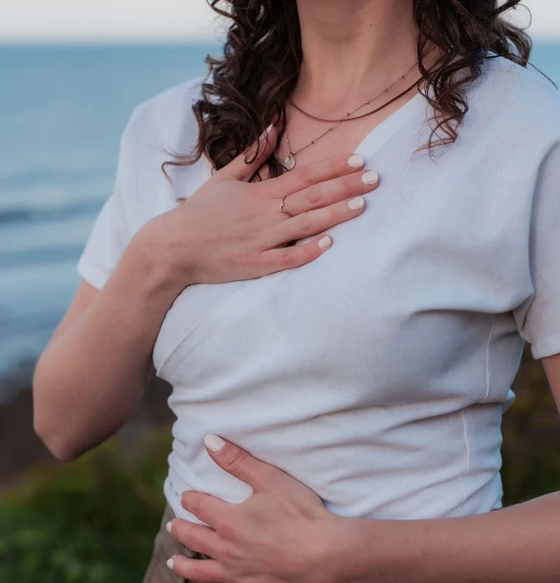 A woman with brown curly hair has one hand on her heart and one on her belly. The background shows the ocean and some greenery.