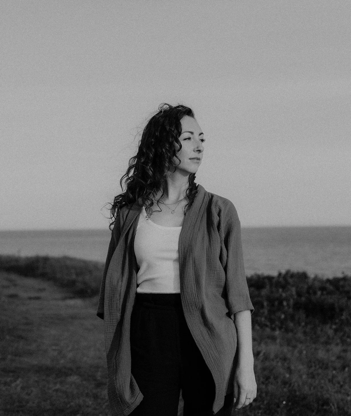 Black & white image of curly-haired woman looking off into the distance. She wears a loose sweater overtop of a white tank top. The background is some grass, ocean and sky.