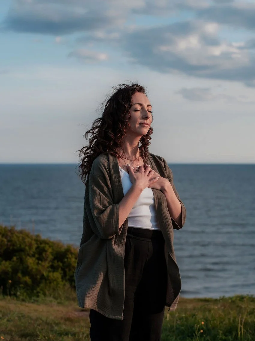 Woman with curly brown hair has hands over heart and eyes closed. She is facing the sun and the ocean is behind her.