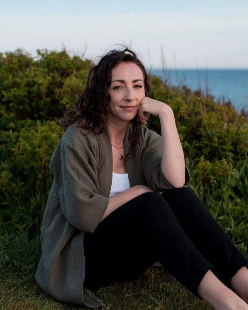A woman with wavy brown hair sitting outdoors on grass near a hedge, near the ocean, smiling gently with her chin resting on her hand, wearing a gray jacket over a white top and black pants.