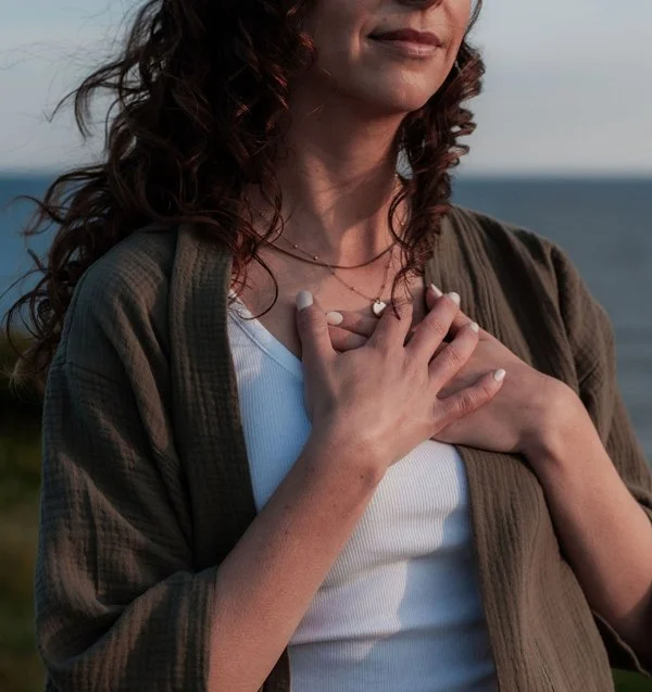 Woman with curly brown hair has her hands over her heart. She wears an olive green sweater over a white top.