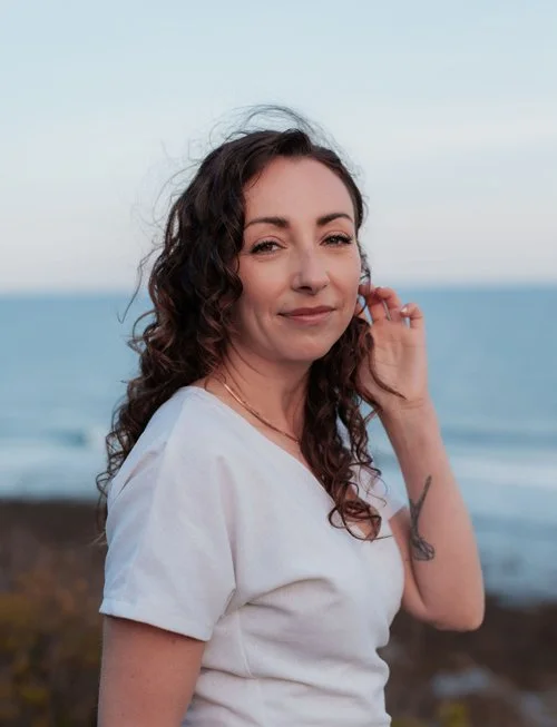 Woman with curly brown hair wearing white shirt is looking at the camera with a smile on her face; she is tucking her hair behind her ear with one hand.